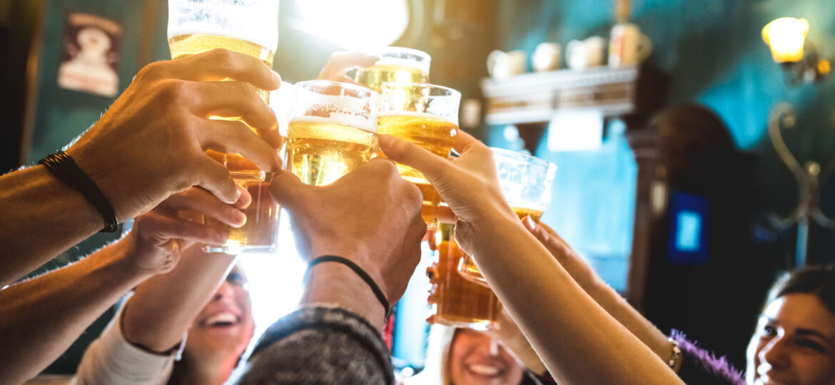 A group of people raising their beer glasses in a celebratory toast inside a warmly lit bar. The scene captures smiles and camaraderie as light shines through the drinks.