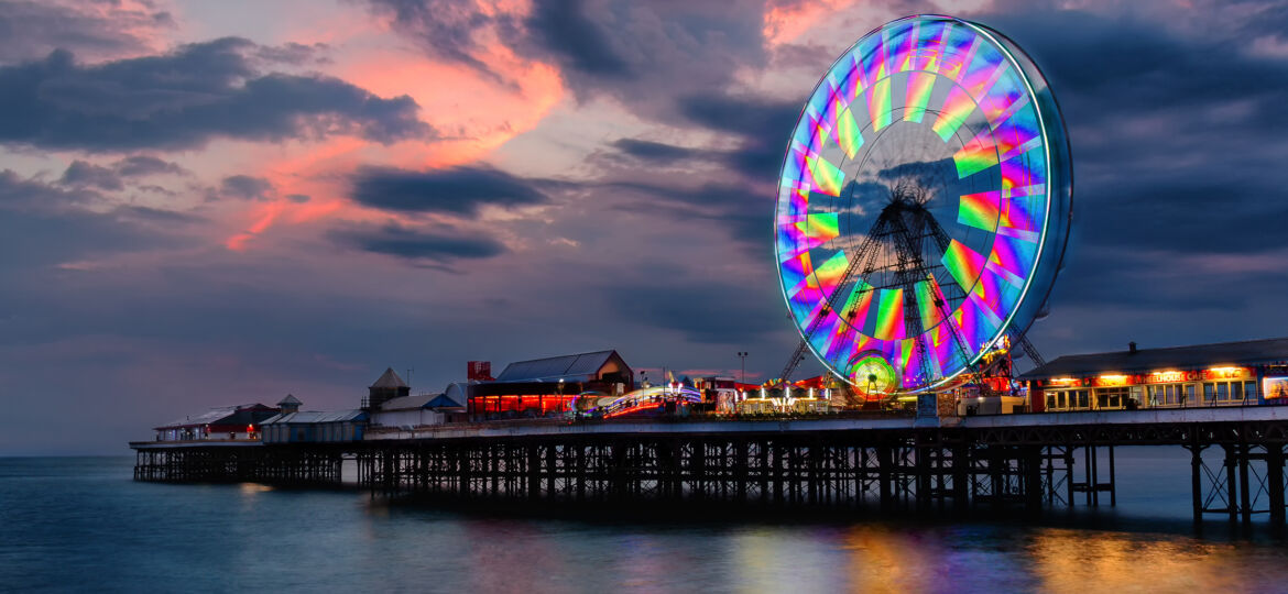 A brightly lit Ferris wheel with vibrant, multicoloured lights on a pier at sunset, reflecting on the calm sea below, under a dramatic sky with dark clouds.