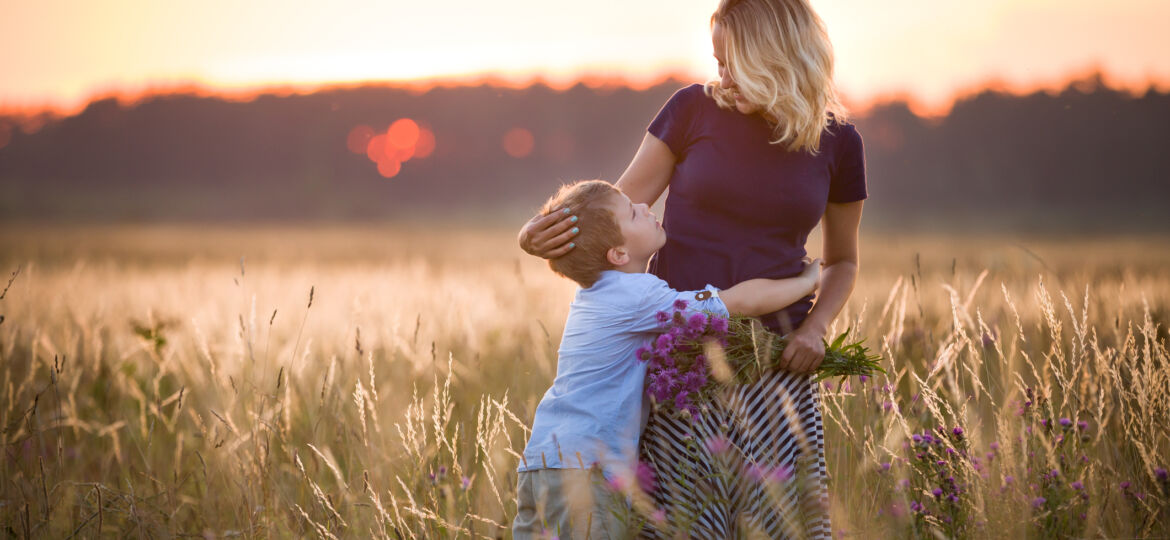 A young boy hugs a woman in a sunlit field at sunset, looking up at her with affection. She holds a bouquet of purple wildflowers while gently touching his head.